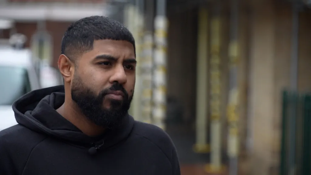Zeshan is standing in an alleyway, wearing a black hooded top. You can see a vehicle behind him and a building with scaffolding outside.