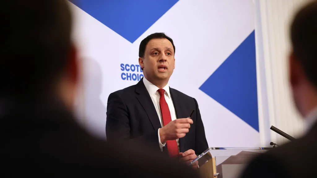 Anas Sarwar on stage at his media conference. He is standing in front of a large Saltire on the wall, faeturing the word Scotland's Choice in blue.