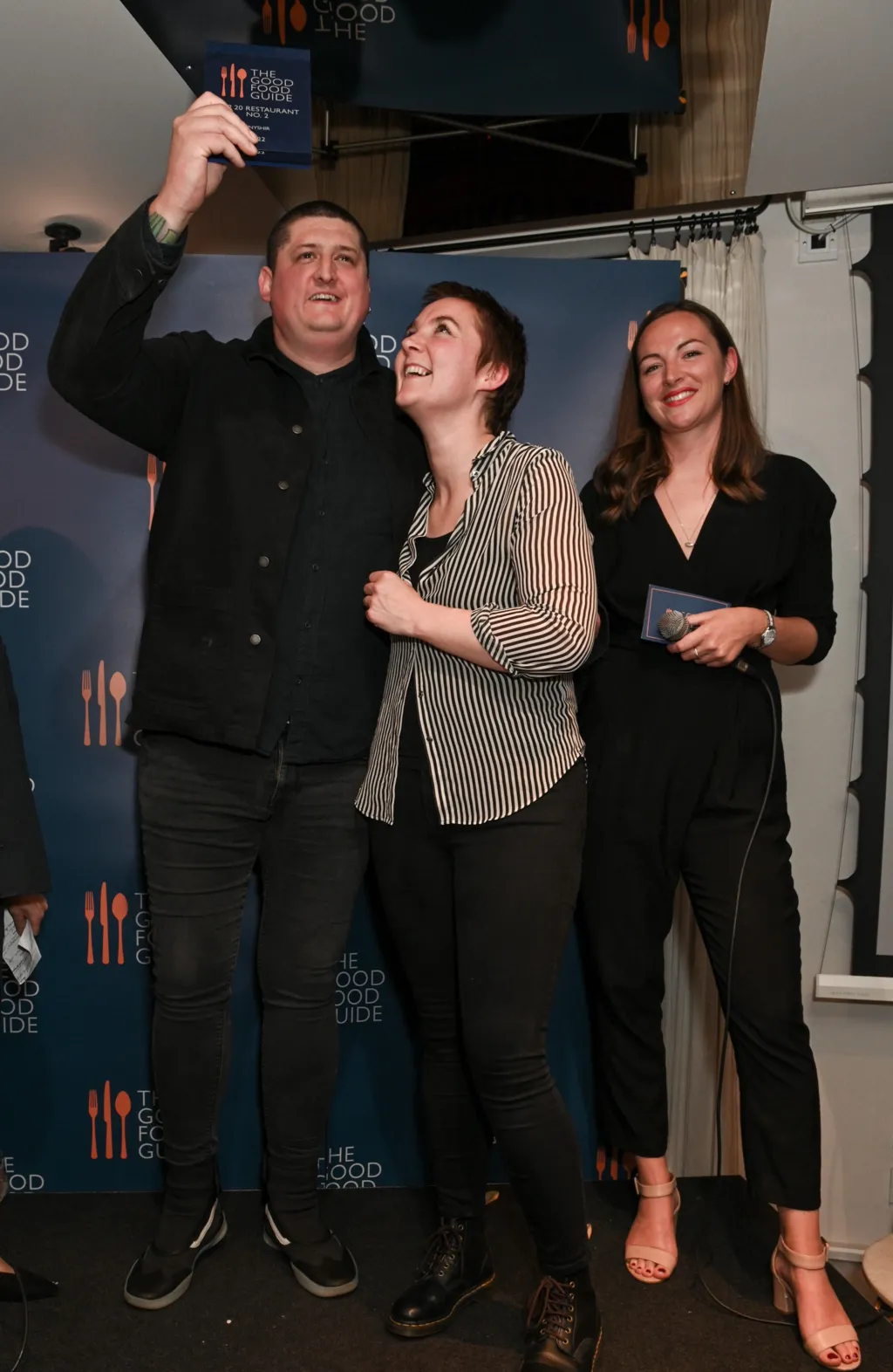 Chef Gareth Ward stands on a small stage in front of a “The Good Food Guide” backdrop. He raises an award plaque above his head while smiling, his guest, a woman, looks up at it, smiling, and a woman on the right smiles while holding a microphone and a card.