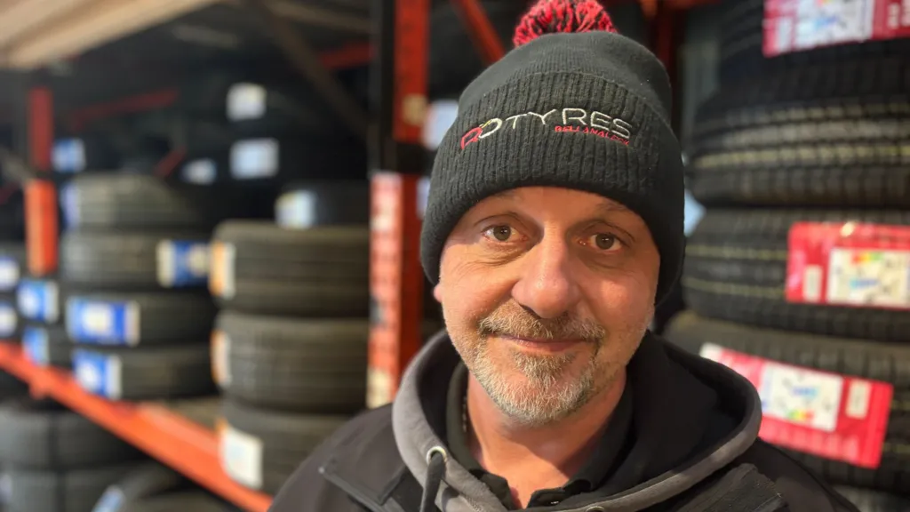 A man stands in a garage with rows of tires on shelves behind him. The man is smiling with a wooly had and warm clothing on. He has a grey beard and is dressed in black and grey but has a red bobble on his hat. 