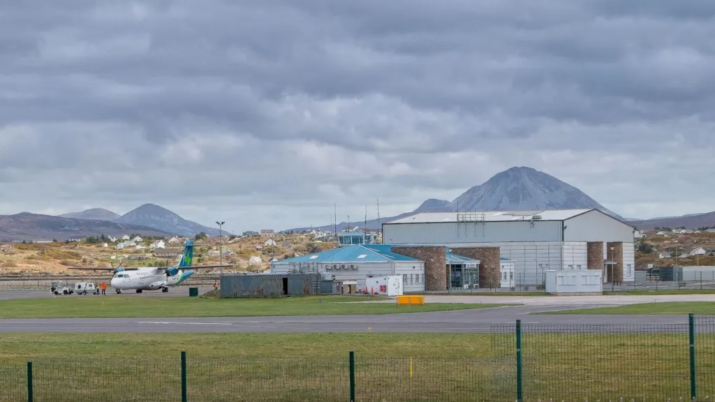  Carrickfinn Airport in County Donegal. An Aer Lingus plane sits on the runway near the airport, a small terminal building with a blue roof is to the right and beside that is a large hangar. Mountains can be seen in the background.
