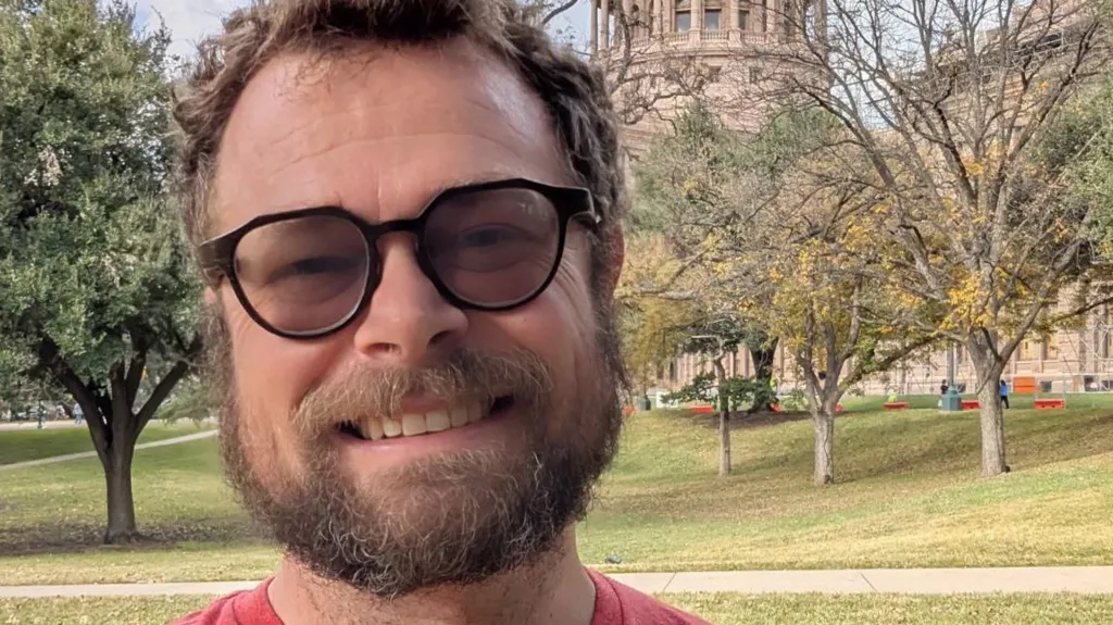 Close up of a man's face with beard moustache and glasses. He is standing outdoors against a backdrop of trees and grass. He is smiling.