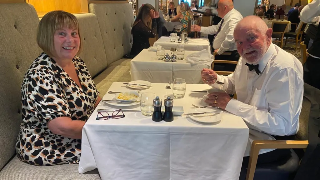 A woman and a man sit at a table in a restaurant, she is wearing an animal print dress and he is in a white w=shirt and black bow tie, and has a white beard.