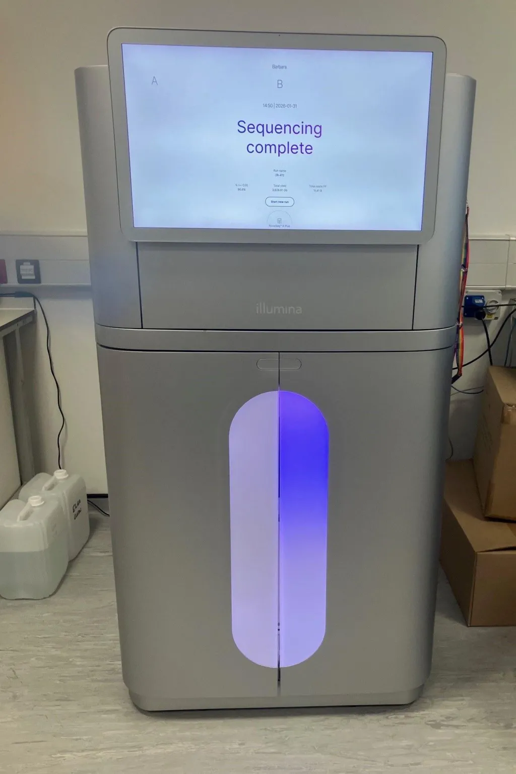 A high-tech silver machine about the height of a person, stands in an office environment. It has a large monitor taking up the top third, showing the words Sequencing Complete. There aren't any controls to be seen, and the bottom half has a purple light going down the middle in an oval shape. 