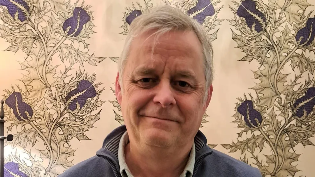 Iain Smith, who has grey hair and is wearing a blue top, stands looking into camera in front of thistle wallpaper 
