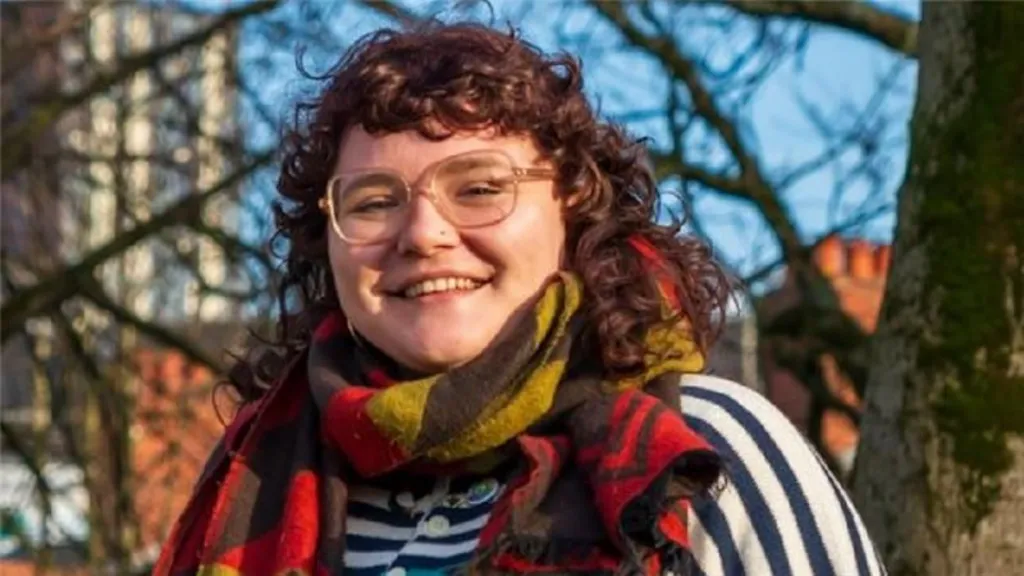 Tessa smiles at the camera. It is a head and shoulders shot of her, she has shoulder length brown curly hair, a nose stud, and wears glasses. She is wearing a yellow, red and black patterned scarf and a blue and white striped top. There are trees behind her, and a sky-scraper in the background. 
