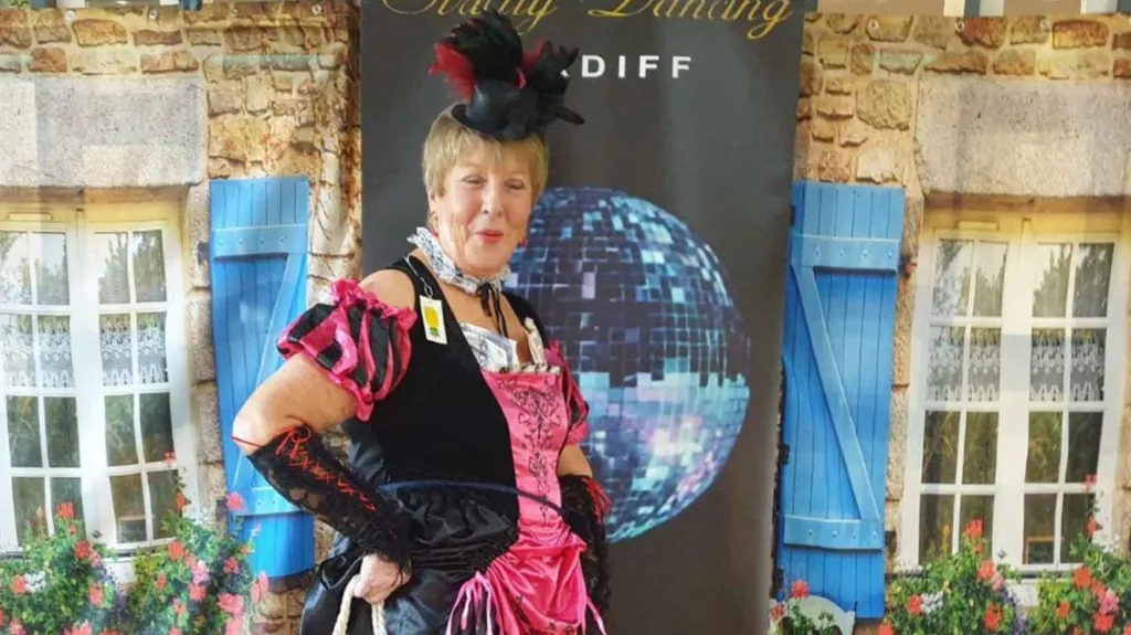 Pat is striking a pose in a flamboyant black and pink ballroom gown, fascinator and pink heels, in front of a popup stand branded with 'Strictly Dancing Cardiff'. 