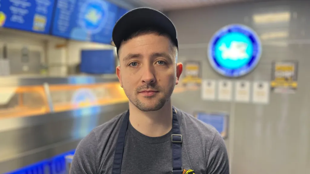 Grant Cranston is standing in Hilton Chip Shop. He is wearing a cap and an apron over a grey T-shirt. Behind him is the shop's counter and menu board.