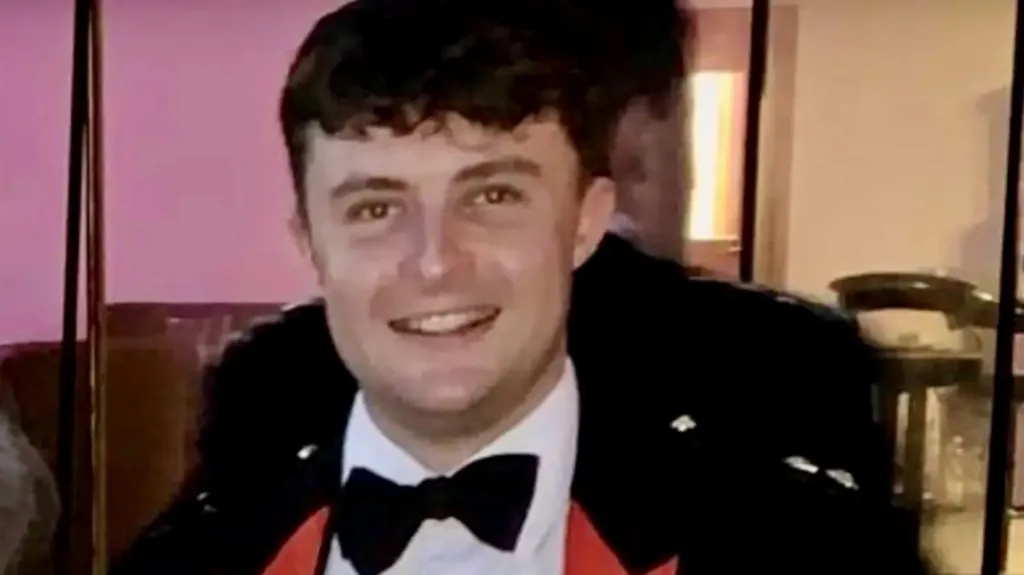 Captain Philip Gilbert Muldowney smiles as he looks into the camera, with short dark hair, wearing a dress suit and bow tie. There is a person standing behind him looking the other way.