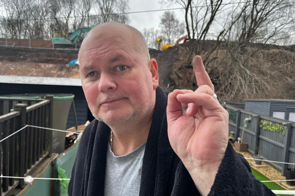 a man showing black soot on his finger standing in front of a building site with diggers on it.