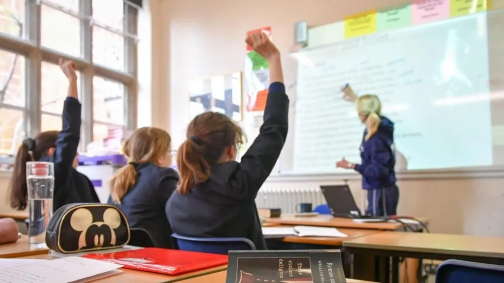 Pupils in a classroom wearing navy uniform sit at desks with their hands up as though about to answer a question. A teacher stands at the front at a whiteboard.