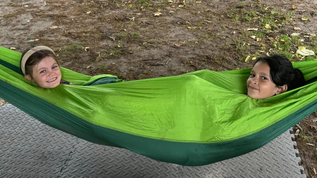 Two children zipped up an a green hammock - only their heads are visible. They are both smiling - one had a headband and the other has dark hair.