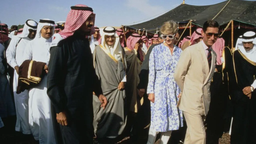 Charles, wearing a cream linen suit and sunglasses, and Princess Diana, wearing a blue and white floral dress and sunglasses, walk along smiling, surrounded by many men in traditional Saudi attire, also smiling.