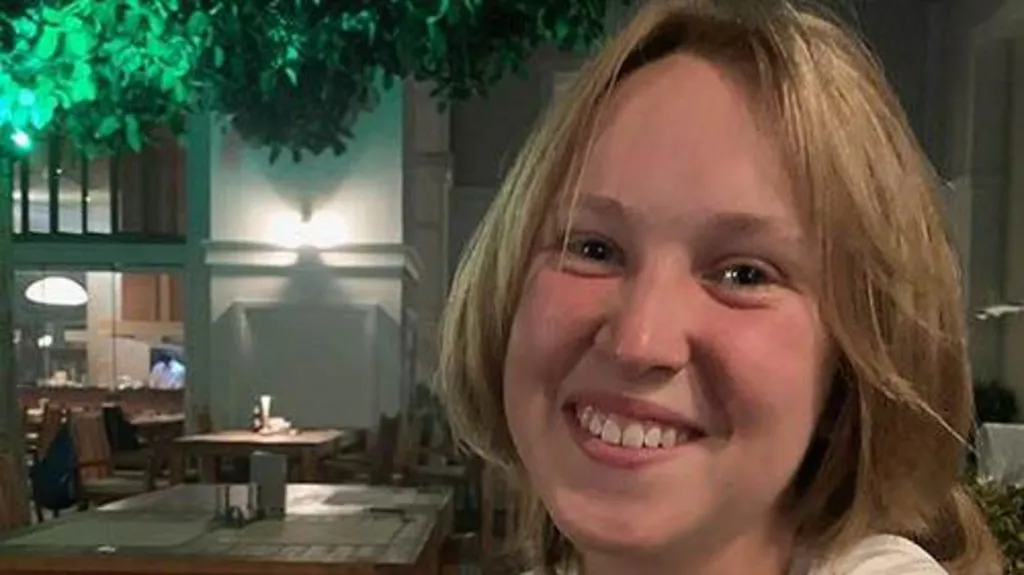 A smiling Georgia looks directly at the camera, she is seated in an empty restaurant. She has short brown hair. 