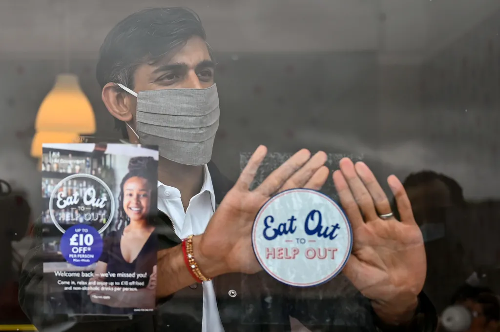 Photograph of Rishi Sunak wearing a mask in the summer of 2020. He is behind a window of a restaurant and attaching a sticker which promotes the Treasury's Eat Out to Help Out scheme.