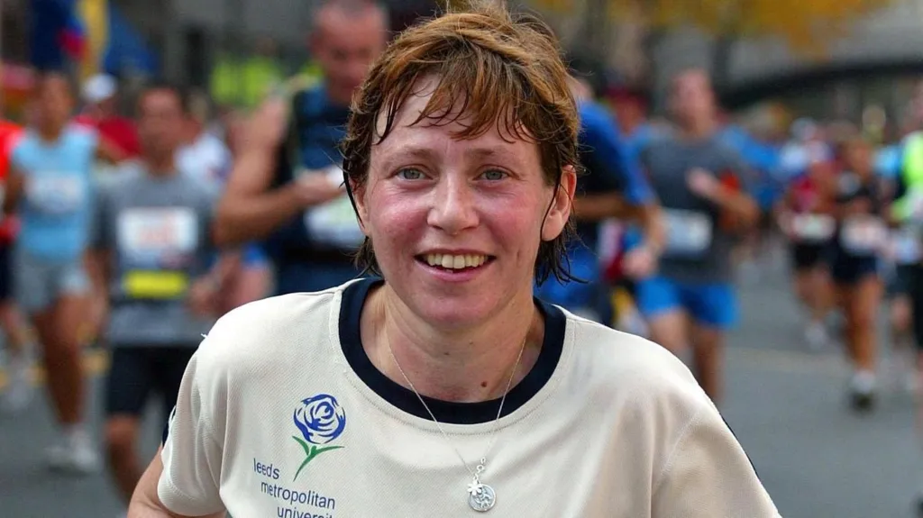 Jane Tomlinson running along a road with many other runners behind her. She is wearing a cream-coloured top marked "Leeds Metropolitan University" and is wearing a necklace with a star. She smiles at the camera.