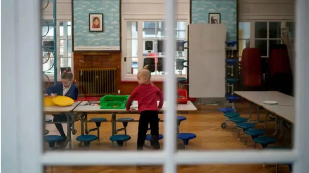 Two children in a school classroom. The boy is wearing red and the girls is wearing blue. The camera is taking the photo from behind a window and the window can be seen in the foreground. 