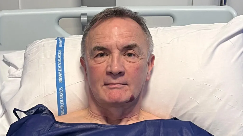 Michael Robinson is in a hospital bed. He is wearing blue patient scrubs and has short white hair. He is resting his head up on a pillow.