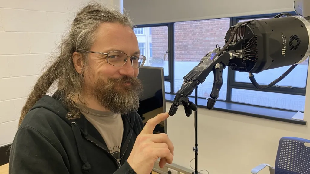 With a beard and long hair Rich Walker smiles at the camera while touching the robot hand.