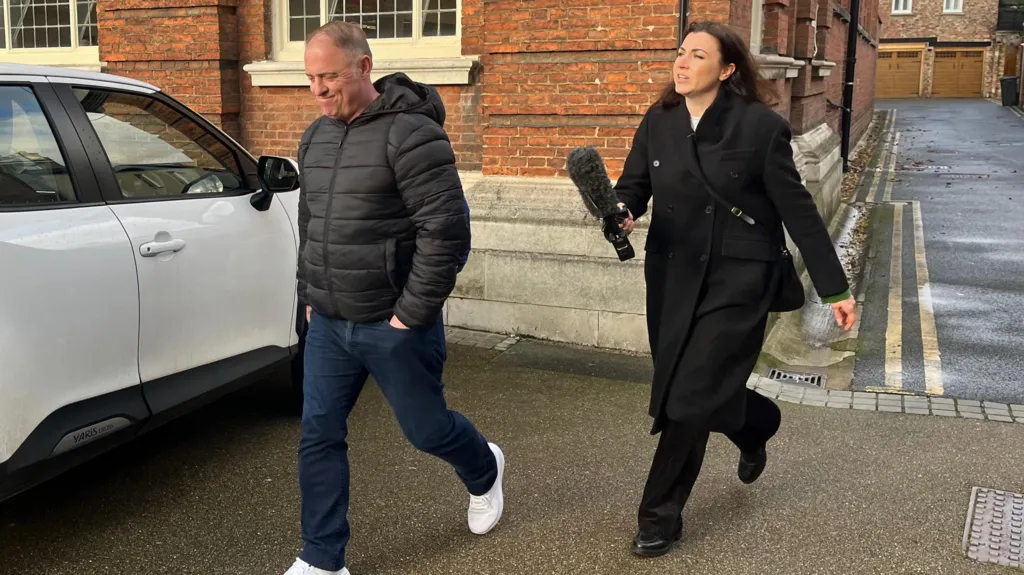 Raymond McDonald, a man in his fifties, is walking away from Philippa Goymer who is a BBC journalist. She is dressed in black and has long dark hair and is carrying a large microphone. He has a black jacket, blue jeans and has his hands in his pockets. Behind them both is a brick built building and a white car is parked to the side.