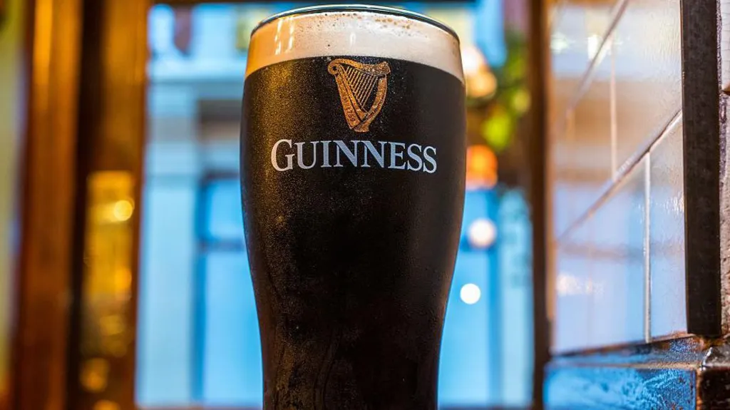 A close up image of a pint of Guinness in a glass depicting the firm's logo. The drink is placed on a bar with a window blurred out in the background.