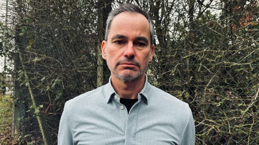 Calvin Jones looks at the camera with a neutral expression. He is in a check shirt in front of a hedge