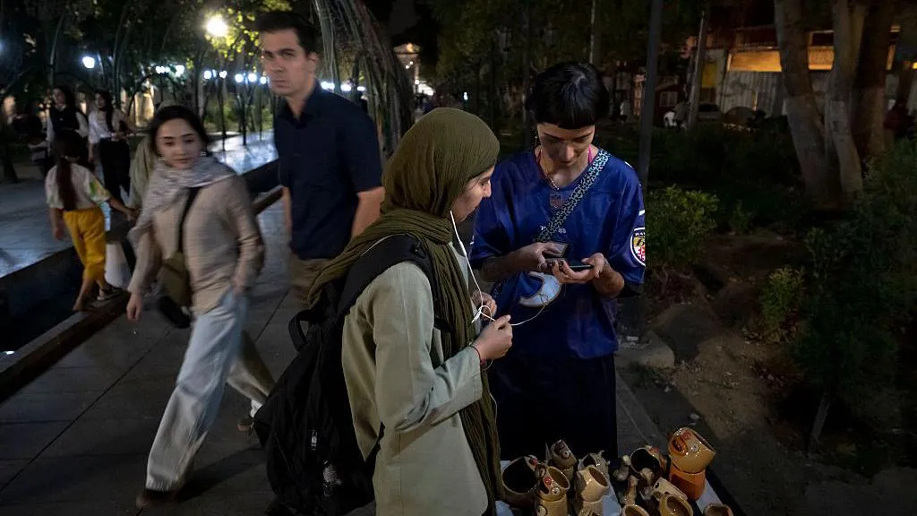 During a nighttime walk in northern Tehran, two young women are standing beside a small display of handmade pottery. One of them is entering information into a customer’s mobile phone. Pedestrians pass by, while the streetlights and surrounding trees illuminate the scene.