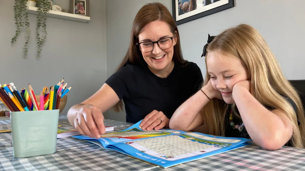 Mum Sara and daughter Ellie are looking at a children's puzzle book on the kitchen table. They are smiling. A pot of coloured pencils is on the table in front of them. 