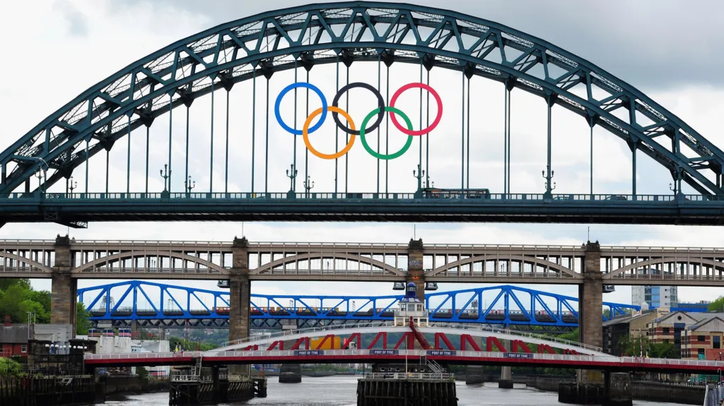 The Olympic Rings logo hangs from the Tyne Bridge. Five colourful, interlocking rings are suspended on the side or an arched metal bridge spanning a river below