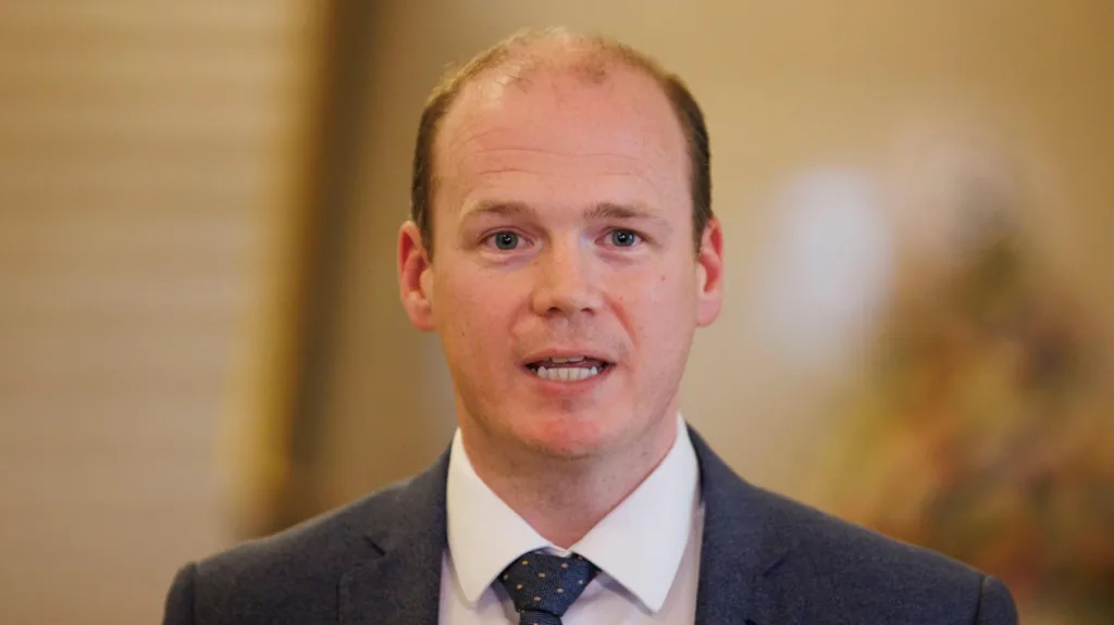Gordon Lyons is standing in front of the camera and looks as though he is speaking. He is wearing a blue jacket with a spotted tie and a white shirt. He has fair brown hair which is thinning on the top