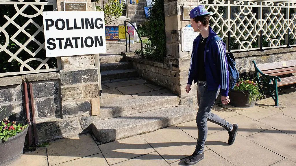 A young man wearing a cap and listening to headphones walks past a sign reading "polling station".