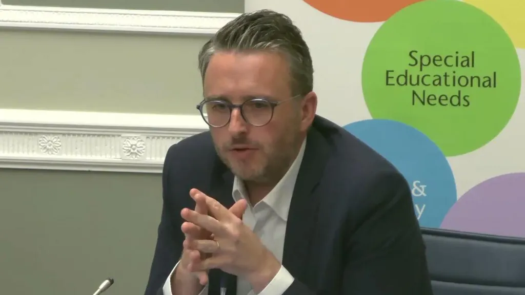 Nick Mathison, a man with short, greying dark hair, sits with his hands clasped during a political meeting.  He is wearing glasses, a white shirt and a navy blazer.  A sign behind his chair reads: "Special Educational Needs". 