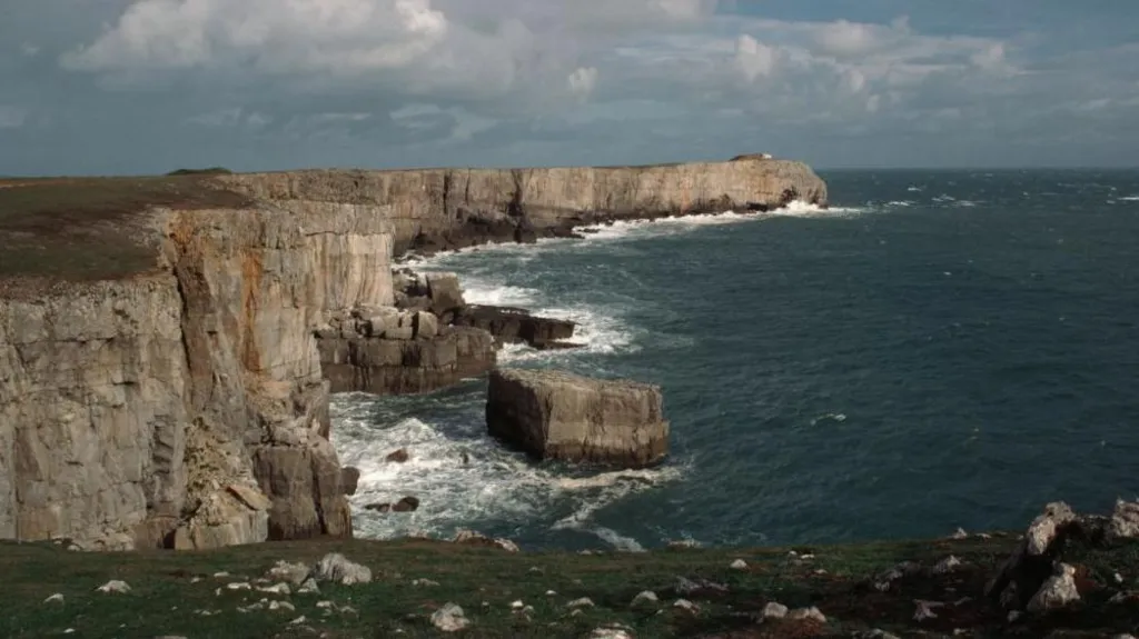 Cliffs at St Govans Head. The sky is blue and cloudy. The waves crash into the cliff face.