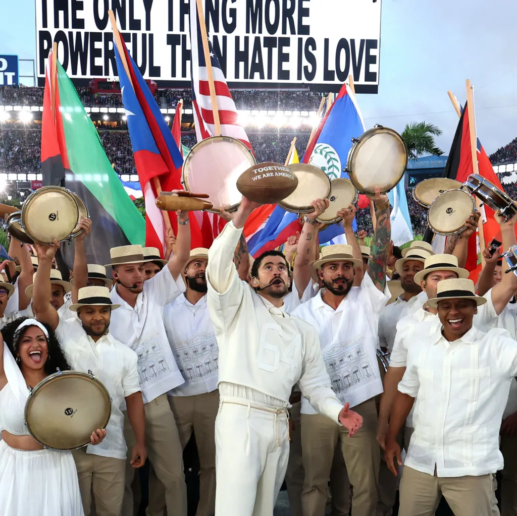 Bad Bunny holding up a football with white writing, surrounded by musicians and dancers holding up instruments and flags