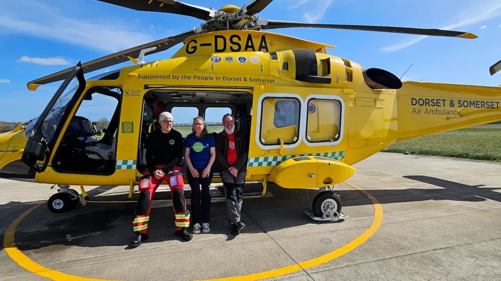 The couple and a member of the air ambulance to their left sitting on the side of an open yellow helicopter. They are all faceing the camera with their hands on their laps and smiling. They are on a concrete surface, with a yellow circle surrounding the helicopter, behind them is green grass.
