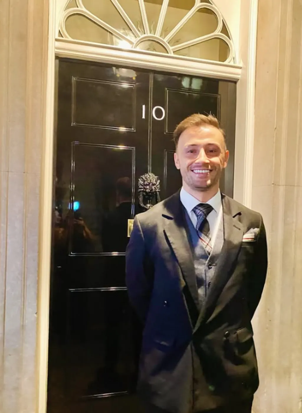 Zander Murray is wearing a dark suit with a tartan tie. He is standing in front of the black door at 10 Downing Street with his hands behind his back while smiling. 