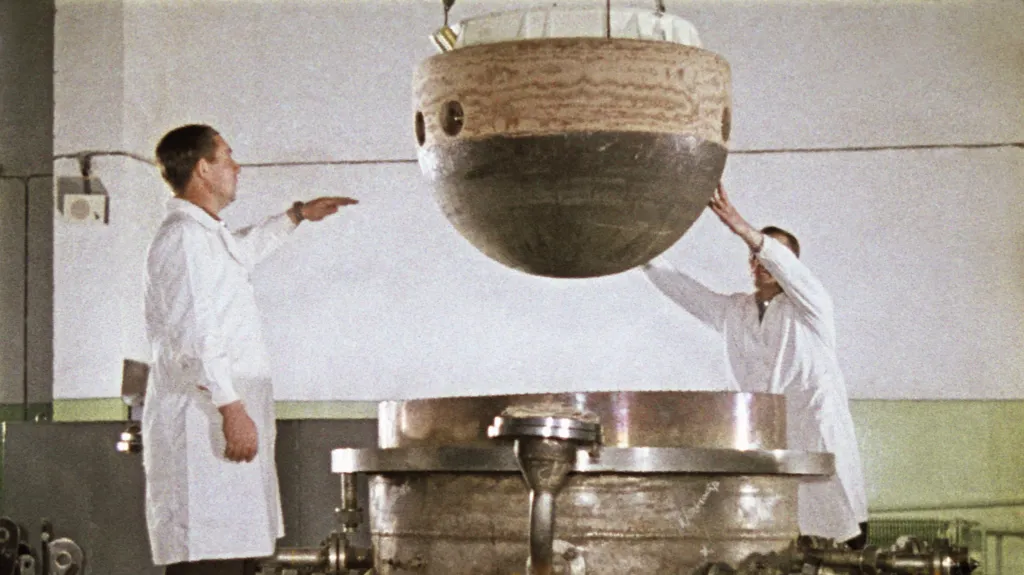 The image shows two individuals in white lab coats lowering the descent capsule of the Soviet space probe Venera 5 or 6 into a large, cylindrical metal structure to test the heat resistance. The capsule is spherical with a dark bottom half and a lighter top half, featuring several circular holes. 