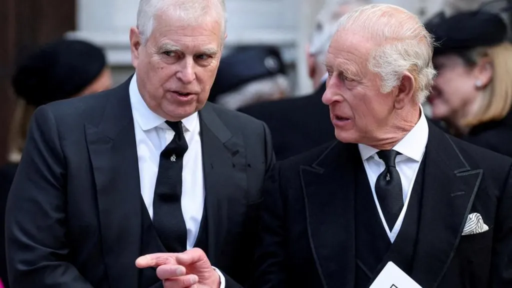 Andrew and King Charles talking as they walk. Charles points across Andrew and looks to the side while Andrew looks ahead. They both wear black suits, white shirts and black ties