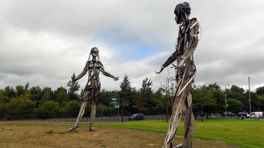 Two semi-abstract steel and bronze statues face eachother at about 18 foot tall, with arms outstretched on a green space. Behind them is a road of traffic, backed by large trees. It is cloudy, interrupted only by a brief glimpse of blue sky in the middle of them.