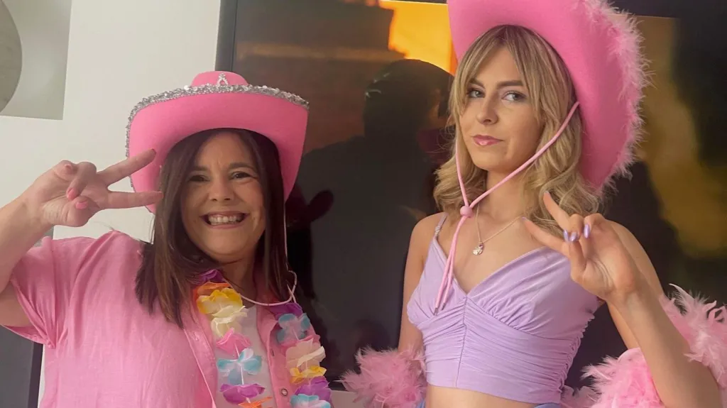 Michelle and Isla Sneddon holding up 'peace' signs and posing for the camera. They are both wearing pink cowboy hats and dressed in pink and purple. Michelle has brown hair and is wearing a flower lei and Isla has blonde hair and is wrapped in a feather boa.