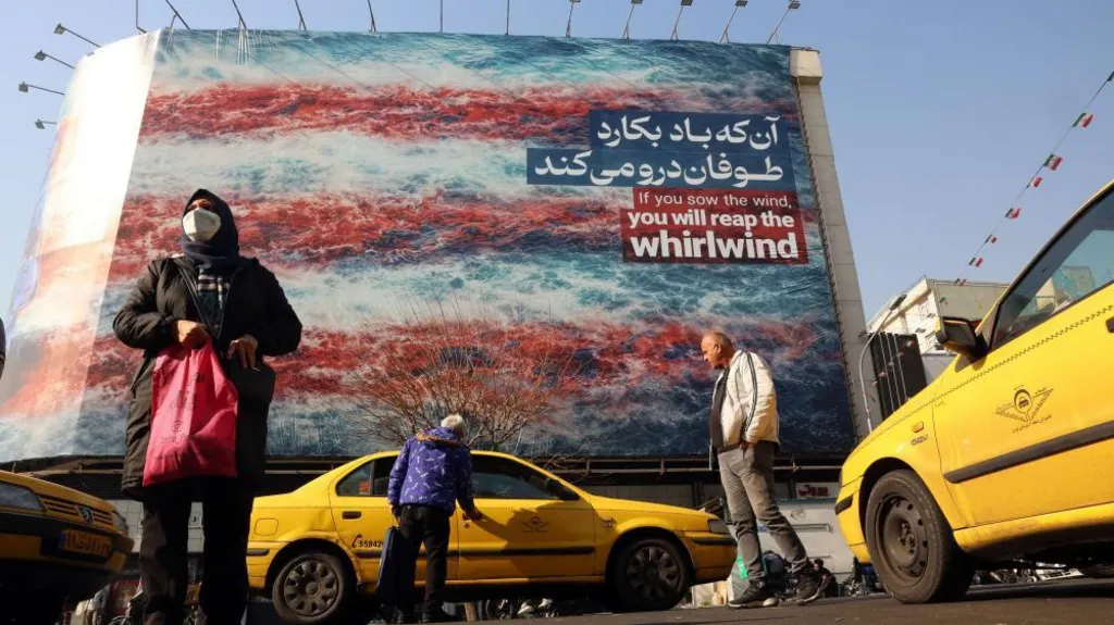 A woman is seen carrying a shopping bag and a man is seen getting into a yellow taxi cab in the foreground of the photo, while in the background there is a huge anti-US billboard which reads 'If you sow the wind, you will reap the whirlwind' 