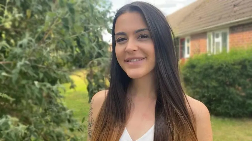 Abbie standing in a garden in front of a house. She has long brown hair and is wearing a white vest top
