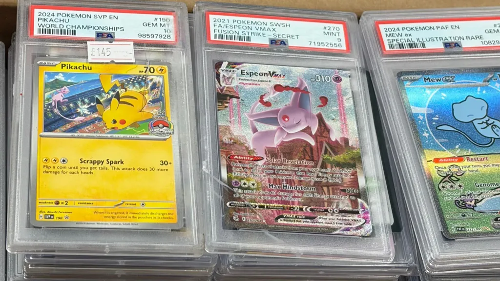 The image shows three damaged Pokémon cards, the clear cases the cards are housed in are cracked and scratched. The card on the left is a yellow Pikachu card with a sticker that says £145 on it, the middle is a pink Espeon card and the card on the left is a blue Mew card, all with cartoon images of the creatures on. 