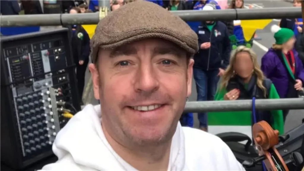 In a selfie, Ben Gunnery is wearing a brown flat cap and a white hoodie. He appears to be on a stage above a crowd with metal railings. There is a black box behind him with various dials and buttons. At the bottom of the photo you can see that he is holding a brown violin. 