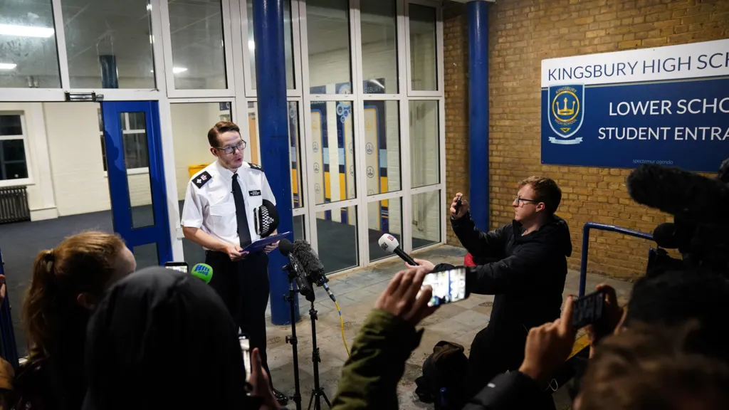 Det Ch Supt Luke Williams gives a statement to reporters outside Kingsbury High School, speaking into microphones as journalists record on phones and cameras near the school’s lower school entrance.