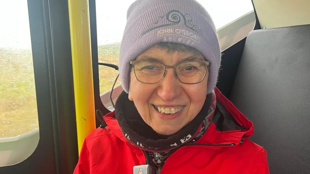 A woman with a purple hat, glasses and a red jacket sitting in the back of an autonomous vehicle