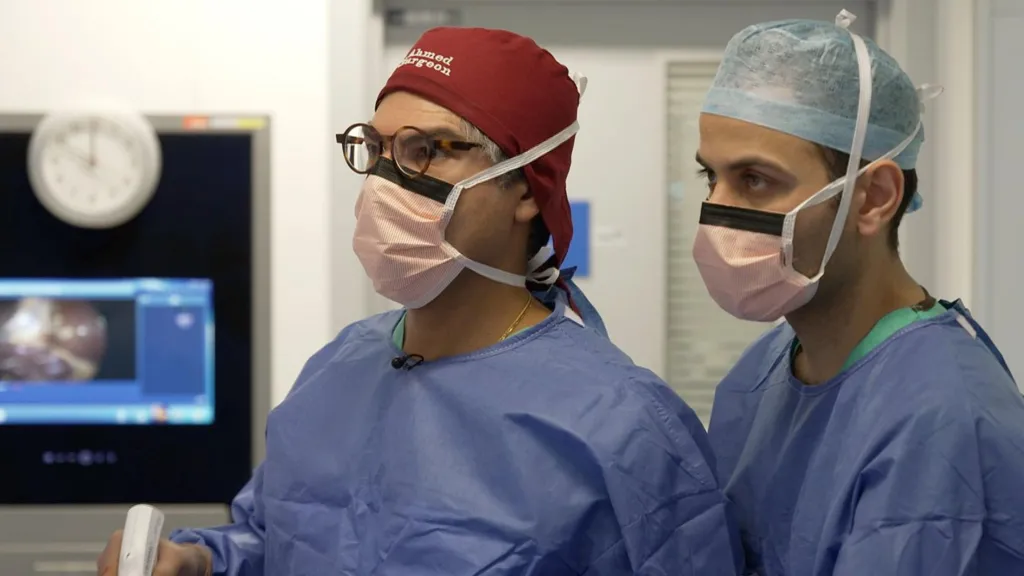 Two male surgeons in blue scrubs and face masks operate on a patient in a hospital. One of the surgeons is holding a white instrument in his hand. Behind them a screen in showing what they are operating on, but this is blurred. 