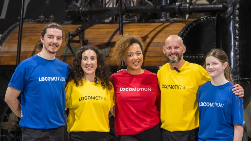 A group of dancers in bright blue, yellow and red t-shirts reading 'Locomotion'. They are stood in front of machinery.
