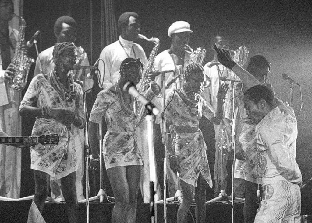 Black and white shot of Fela Kuti with his back to the audience and facing his back-up singers and band. He is singing with one hand up and finger pointing and the other hand behind his back in a pose like a torero.