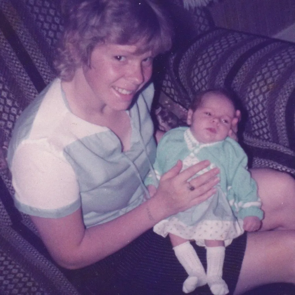 Cathy sits in an armchair with baby Kirsty. Cathy's hair is short and she wears a short sleeved green and white top and a dark skirt. Kirsty is dressed in a white spotted dress with a green cardigan over it and white knee length socks.